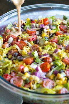 a salad in a glass bowl with a wooden spoon and dressing being poured into it