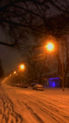 a snowy street at night with cars parked on the side and lights in the background