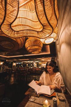 a woman sitting at a table with a book in front of her and lights hanging from the ceiling