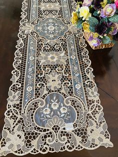 a table runner with white lace and flowers in the background on a wooden table top