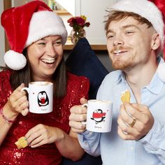 a man and woman sitting on a couch holding coffee mugs with santa hats on them