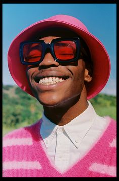 a man wearing sunglasses and a pink sweater smiles at the camera while standing in a field