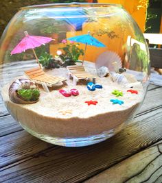 a fish bowl filled with sand, shells and umbrellas on top of a wooden table