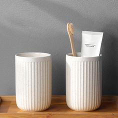 two white cups with toothbrushes in them sitting on a wooden table next to a gray wall