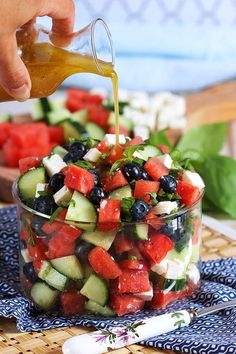 a person pouring dressing into a bowl filled with cucumbers, tomatoes and blueberries