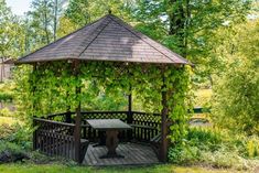 a wooden gazebo surrounded by greenery in the middle of a park with a picnic table underneath it