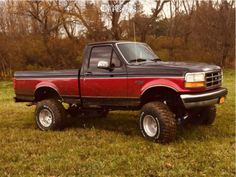 a red pick up truck parked on top of a lush green field with trees in the background