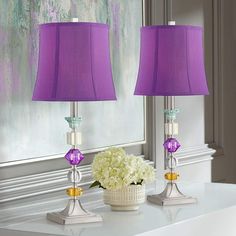 two purple lamps sitting next to each other on a white table with flowers in vases