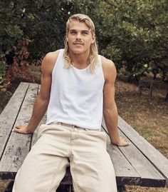 a young man sitting on top of a wooden bench in front of trees and bushes