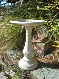 a white bird bath sitting on top of a cement slab in front of some plants