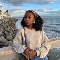 a young woman leaning on a wooden fence near the ocean with her hands in her pockets