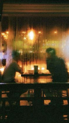 two people are sitting at a table in front of a window with lights on it