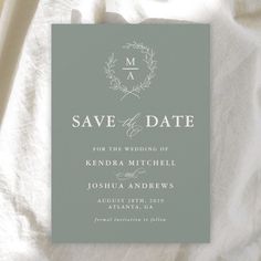 a wedding save the date card on top of a white sheet with an olive wreath