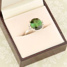 A stunning second hand 18ct white gold green tourmaline & diamond dress ring. An exquisite deep green tourmaline sits at the centre of this gorgeous ring, flanked by a duo of petite diamonds each side. The simple yet stylish setting shows the centre stage gemstone off magnificently whilst the diamonds compliment it perfectly. A part of our second hand gold ring collection. SIZE UK N ½ / US 6 ¾ SPECIFICATION Tourmaline carat weight approx. 5 carats Face dimensions. 11.5 x 14.5 mm Weight. 5.3 gram Elegant Green Topaz Ring In Sterling Silver, Elegant Green Topaz Promise Ring, Timeless Green Wedding Dress For Formal Occasion, Elegant Tourmaline Diamond Ring With Accent Stones, Timeless Green Sterling Silver Ring, Elegant Tourmaline Wedding Ring, Elegant Wedding Tourmaline Rings, Elegant Oval Tourmaline Emerald Ring, Elegant Tourmaline Ring With Center Stone