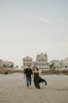 a man and woman holding hands walking on the beach with houses in the background at sunset