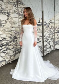 a woman standing in front of a marble wall wearing a white wedding dress with long sleeves