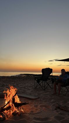 a man sitting in a chair next to a campfire on the beach at sunset