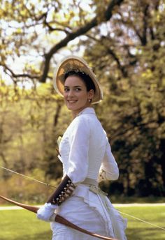 a woman in a white dress and hat is holding a bow while walking through the grass