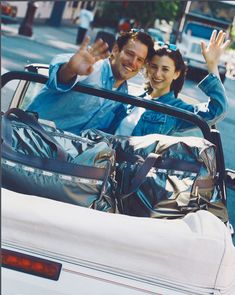 a man and woman waving from the back of a convertible car with luggage in it