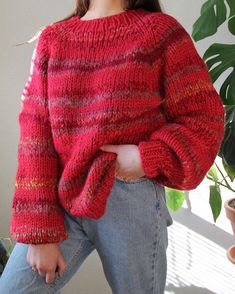 a woman wearing a red sweater and jeans standing next to a potted plant with her hands in her pockets