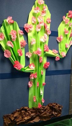 a cactus made out of green paper with pink flowers on the top and bottom, in front of a blue wall