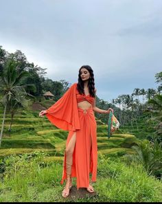 a woman in an orange dress standing on the side of a lush green field with trees