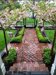 a brick pathway with trees and flowers in the background, leading to a gated area