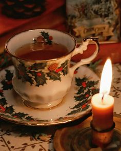 a lit candle sits next to a teacup and saucer with holly designs on it