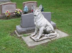 a statue of a dog sitting on top of a cement slab in the grass next to a headstone