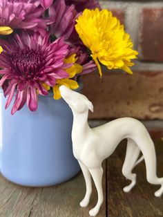 a white dog figurine standing next to a blue vase filled with yellow and purple flowers