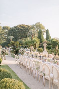 a long table set up with white chairs and flowers in the center is surrounded by greenery