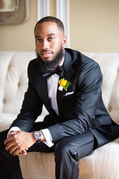 a man in a tuxedo sitting on a couch wearing a flower boutonniere