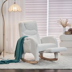 a white rocking chair with a blanket on it in front of a window covered by blinds