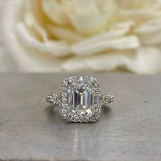 an engagement ring with a large diamond center surrounded by smaller diamonds on a table next to a white rose