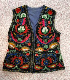 This gorgeous vest features vibrant embroidery in a botanical and floral motif. There are shades of blue, green, red, yellow, pink, and white on a black background. This is a lightweight vest that is perfect for almost any season, and the interior is lined with a soft denim colored fabric. In fact, this vest could be worn in reverse if you wanted a versatile piece. I would describe the condition of this stunning piece as very good, with some minor wear and pilling, especially along the edges, as Folk Style Embroidered Vest For Spring, Festive Folk Vest With Floral Embroidery, Folk Style Cotton Vest With Floral Embroidery, Folk Style Vest With Multicolor Floral Embroidery, Bohemian Multicolor Floral Embroidered Vest, Embroidered Vest, Folk Floral, Lightweight Vest, Colored Fabric