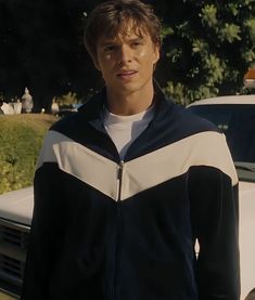 a young man standing next to a white car in front of some bushes and trees