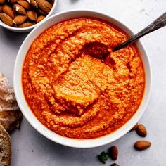 two bowls filled with red sauce next to bread and almonds on a white surface