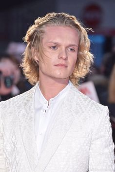 a man with blonde hair wearing a white suit and looking at the camera while standing in front of a crowd