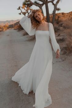 a woman in a white dress is standing on the dirt road with her arms behind her head