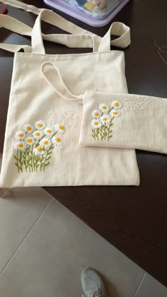 two white bags with daisies on them sitting next to a pair of silver shoes