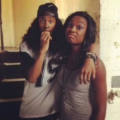 two young women standing next to each other in front of a wall and looking at the camera