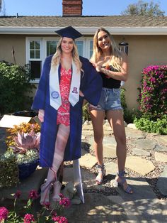 two women standing in front of a house wearing graduation gowns and holding bouquets