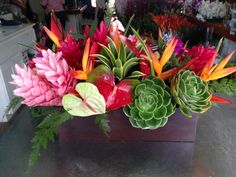 an arrangement of colorful flowers in a wooden vase on a metal table with people standing behind it