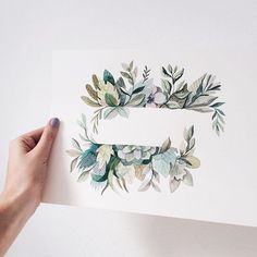 a person holding up a piece of paper with watercolor leaves on it and the letter e