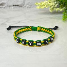 a green and yellow bracelet with the word neymar on it, sitting next to a plant