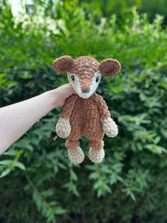 a hand holding a small stuffed animal in front of some bushes