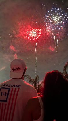 two people looking at fireworks in the sky with palm trees behind them and one person wearing a white hat