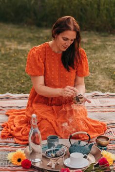 a woman in an orange dress is sitting on a blanket with tea and flowers around her