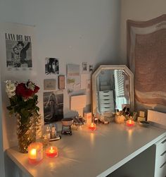 a white desk topped with lots of candles next to a vase filled with flowers and pictures