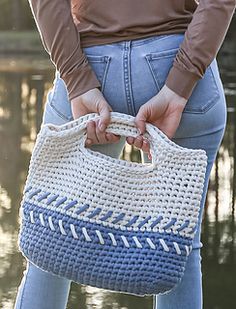 a woman is holding a crocheted bag in her hands while standing by the water